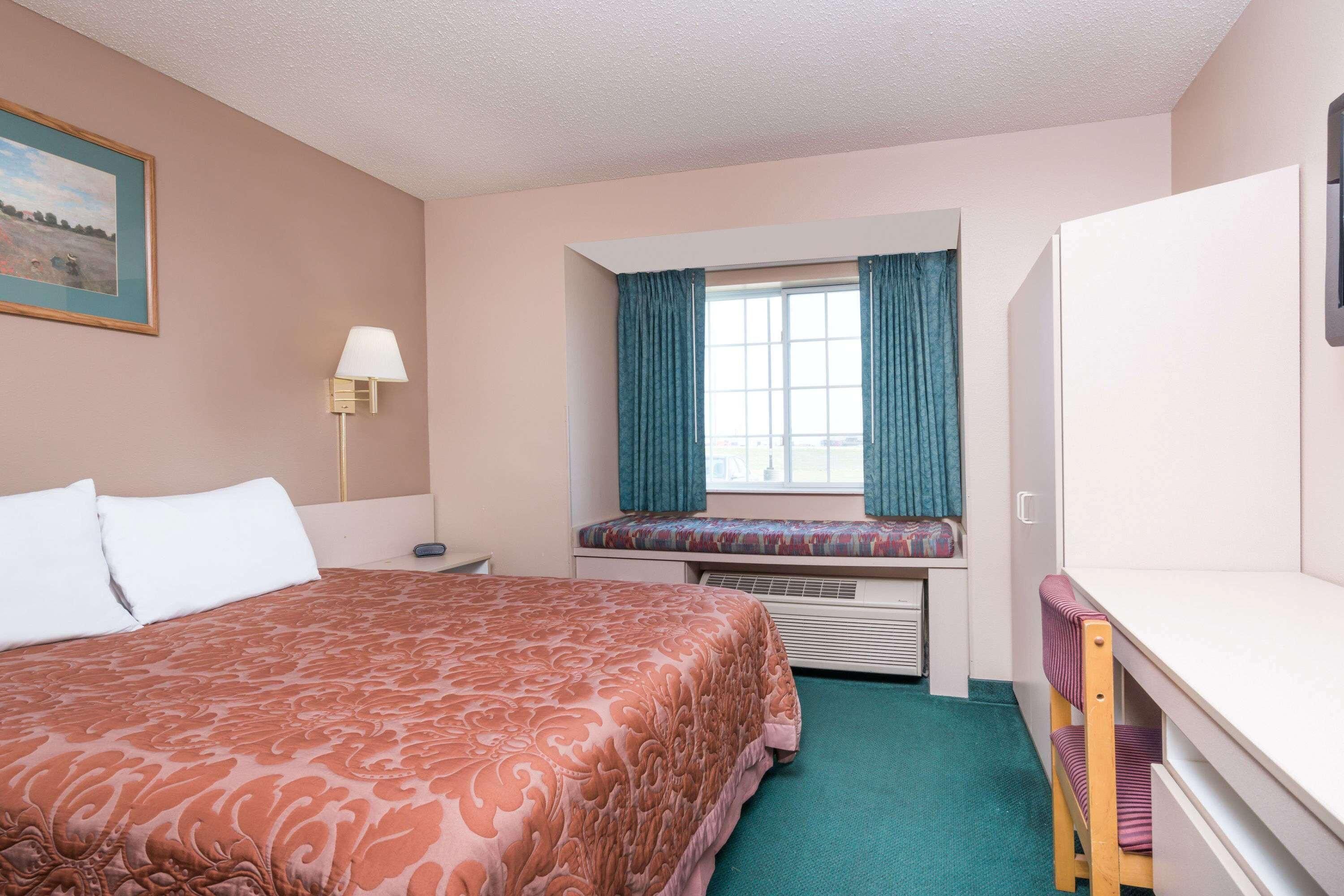Une chambre d’hôtel avec un grand lit, un bureau, une chaise, une fenêtre avec des rideaux, une lampe et un tableau au mur.