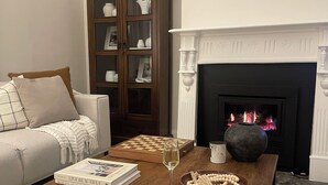 Een gezellige woonkamer met een open haard, een bank, een bijzettafeltje met boeken en een glas wijn, en een decoratieve schaal.