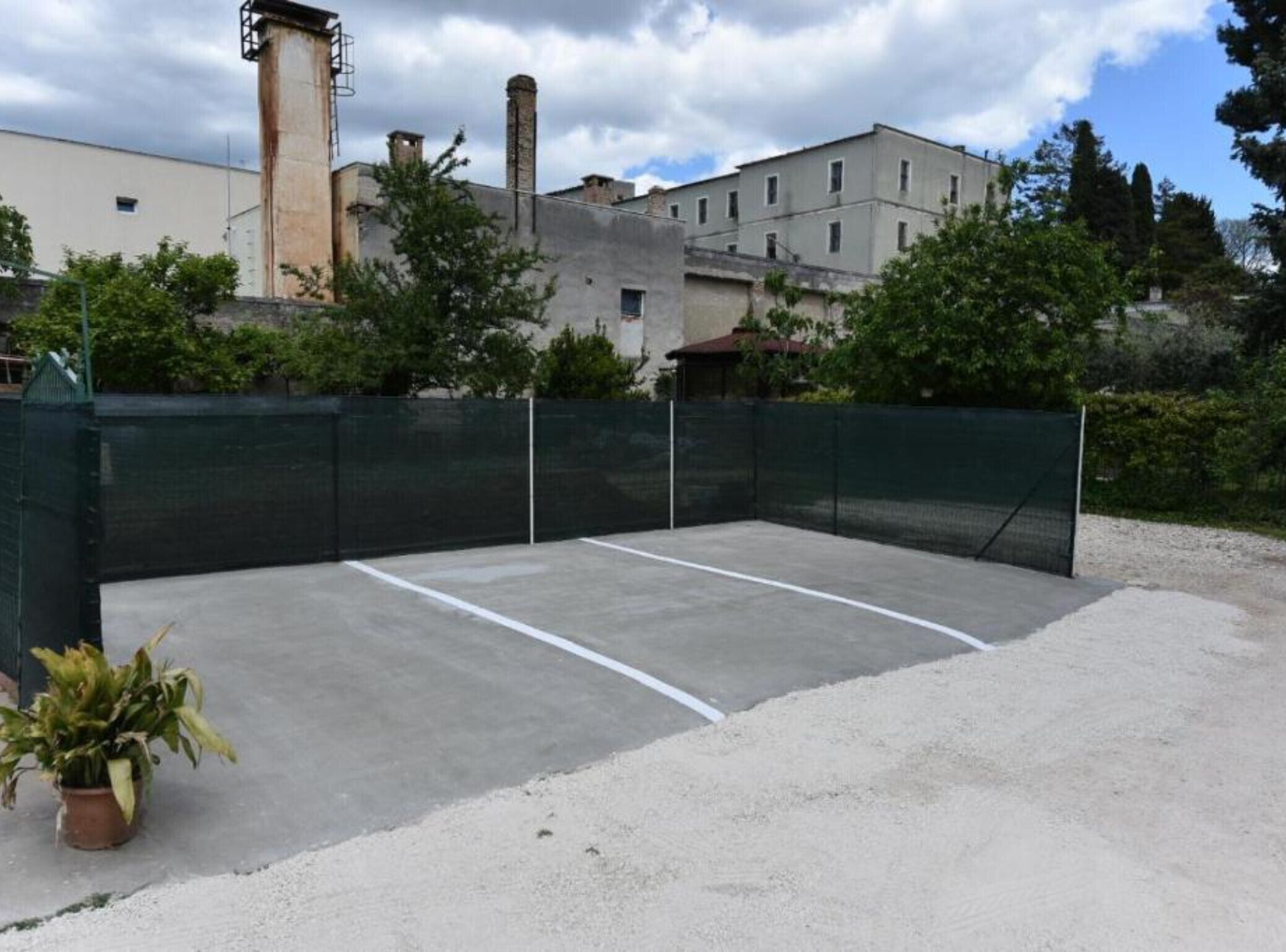 An outdoor tennis court with a net, surrounded by a green fence, and buildings in the background.