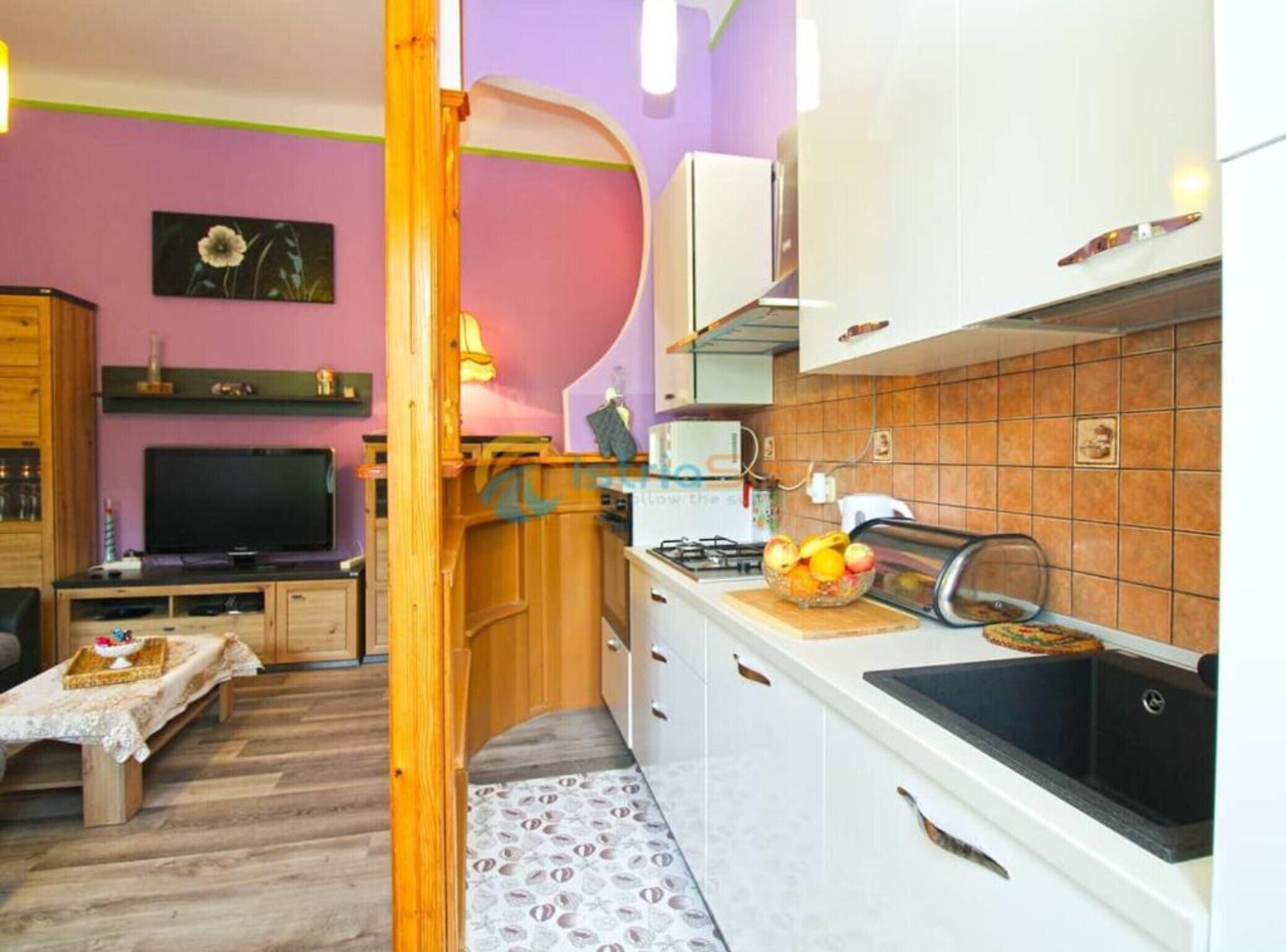 Une cuisine compacte équipée d’une cuisinière à gaz, d’un évier et d’un plan de travail sur lequel se trouve un panier de fruits. On y trouve également une table à manger en bois avec un banc, un téléviseur et une étagère murale agrémentée d’objets décoratifs.