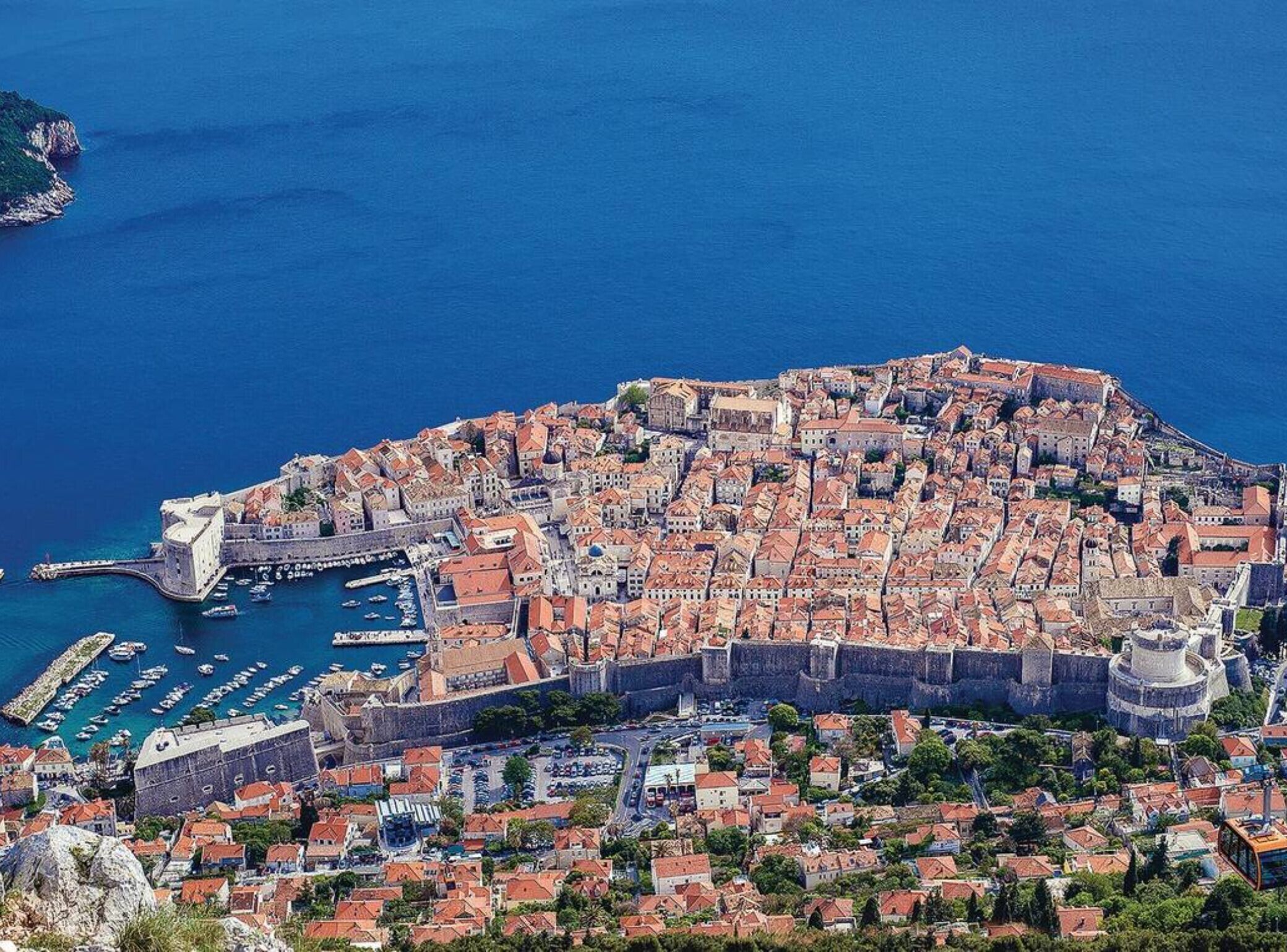 Vue aérienne d’une ville côtière historique, avec ses remparts, un port où sont amarrés des bateaux, et un mélange de bâtiments traditionnels et modernes.