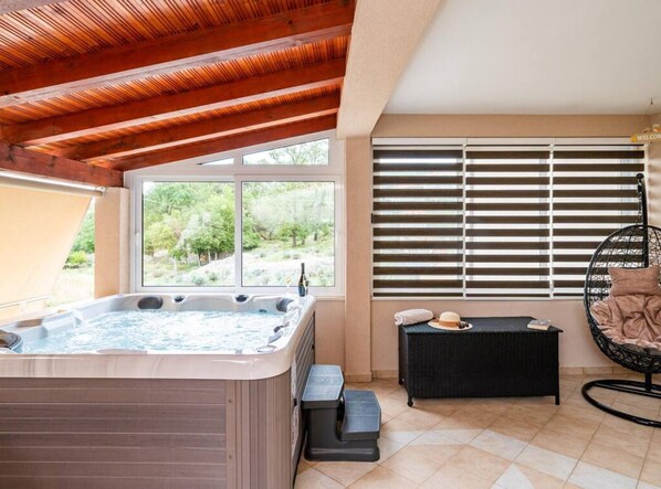 A hot tub in a covered outdoor area with a view of greenery, a wicker sofa, and a hanging chair.