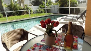 A glass table with a floral tablecloth, a vase with red flowers, a bottle, and two glasses.