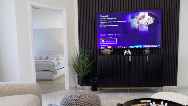 A modern living room with a TV displaying a cooking show, a sofa, a decorative tray, and a plant.