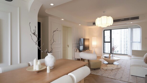 A modern living room with a wooden dining table, white chairs, a white sofa, a TV, and a large window with a view of buildings.