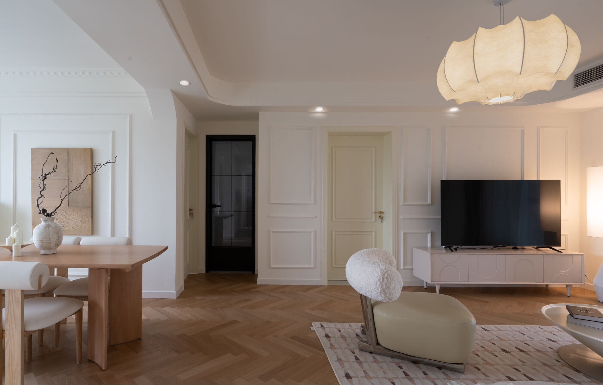 A modern living room with a wooden floor, a dining area with a table and chairs, a white sofa, a TV on a cabinet, and a unique ceiling light fixture.