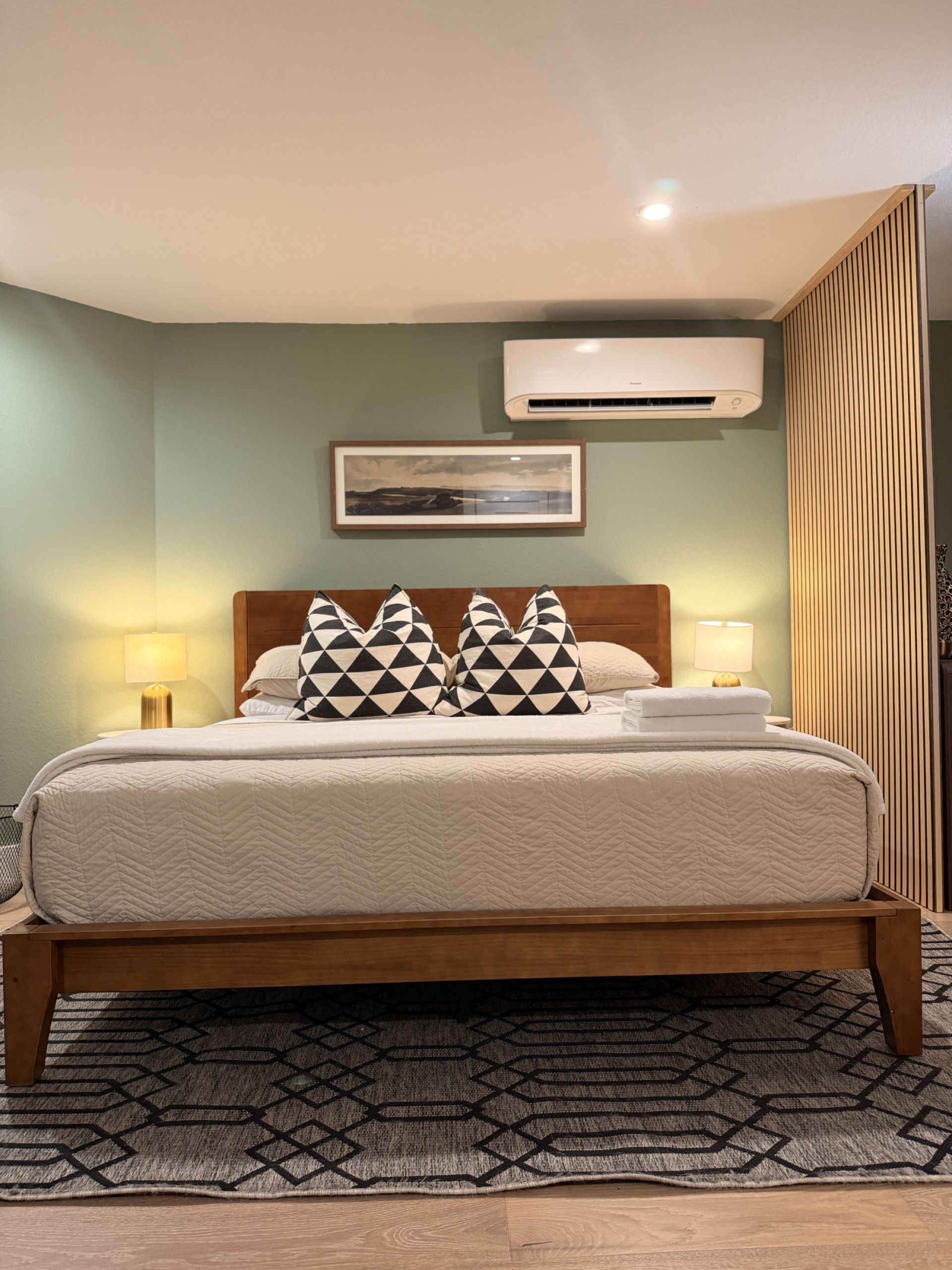 A neatly made bed with a geometric patterned rug, a framed picture on the wall, and an air conditioning unit above it.