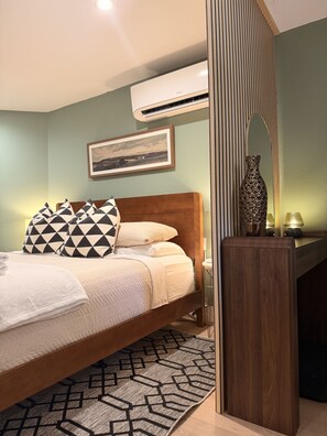 A neatly arranged bedroom with a bed, pillows, a bedside table with a lamp, a mirror, and an air conditioning unit on the wall. - Private King Studio | Near Domain & Apple (Austin)