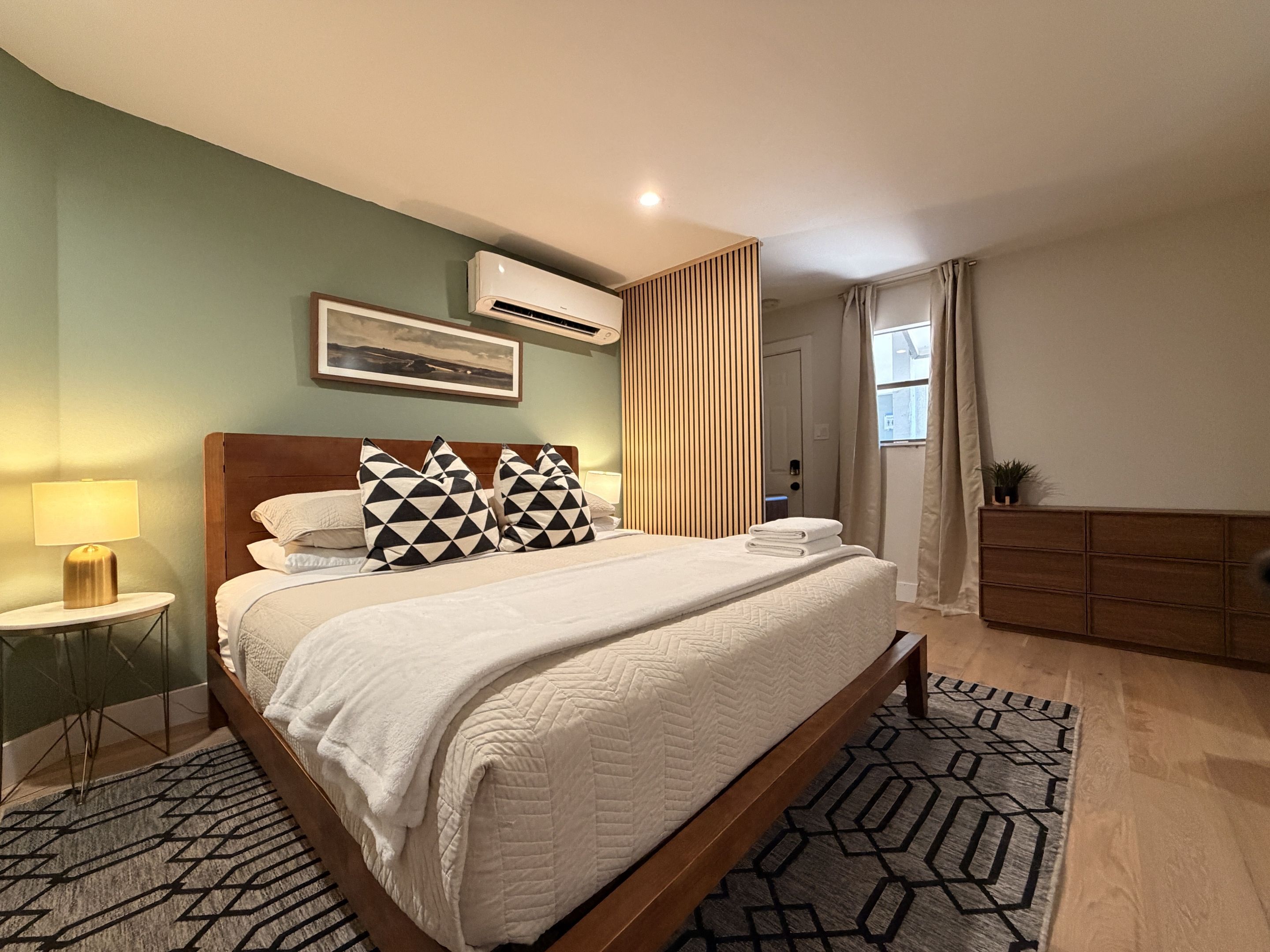 A hotel room with a bed, bedside tables, a wall-mounted air conditioner, a window with curtains, and a small potted plant.