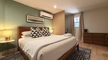 A hotel room with a bed, bedside tables, a wall-mounted air conditioner, a window with curtains, and a small potted plant.