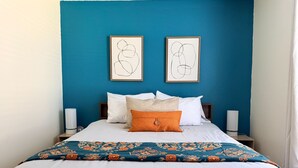 Een bed met wit beddengoed, een blauwe wand en twee ingelijste abstracte kunstwerken.