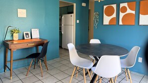 Een moderne eethoek met een ronde tafel, witte stoelen, een houten bureau en een koelkast op de achtergrond.