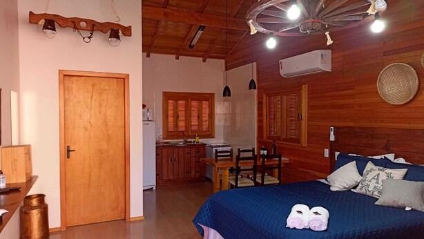 A cozy room with a wooden ceiling, a bed with a blue bedspread, a dining table with chairs, and a kitchen area with wooden cabinets and a window with shutters. - Aconchego da Serra Geminados (São Joaquim)