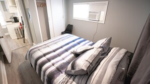 A bedroom with a bed, a chair, a window with an air conditioning unit, and a door leading to a bathroom.