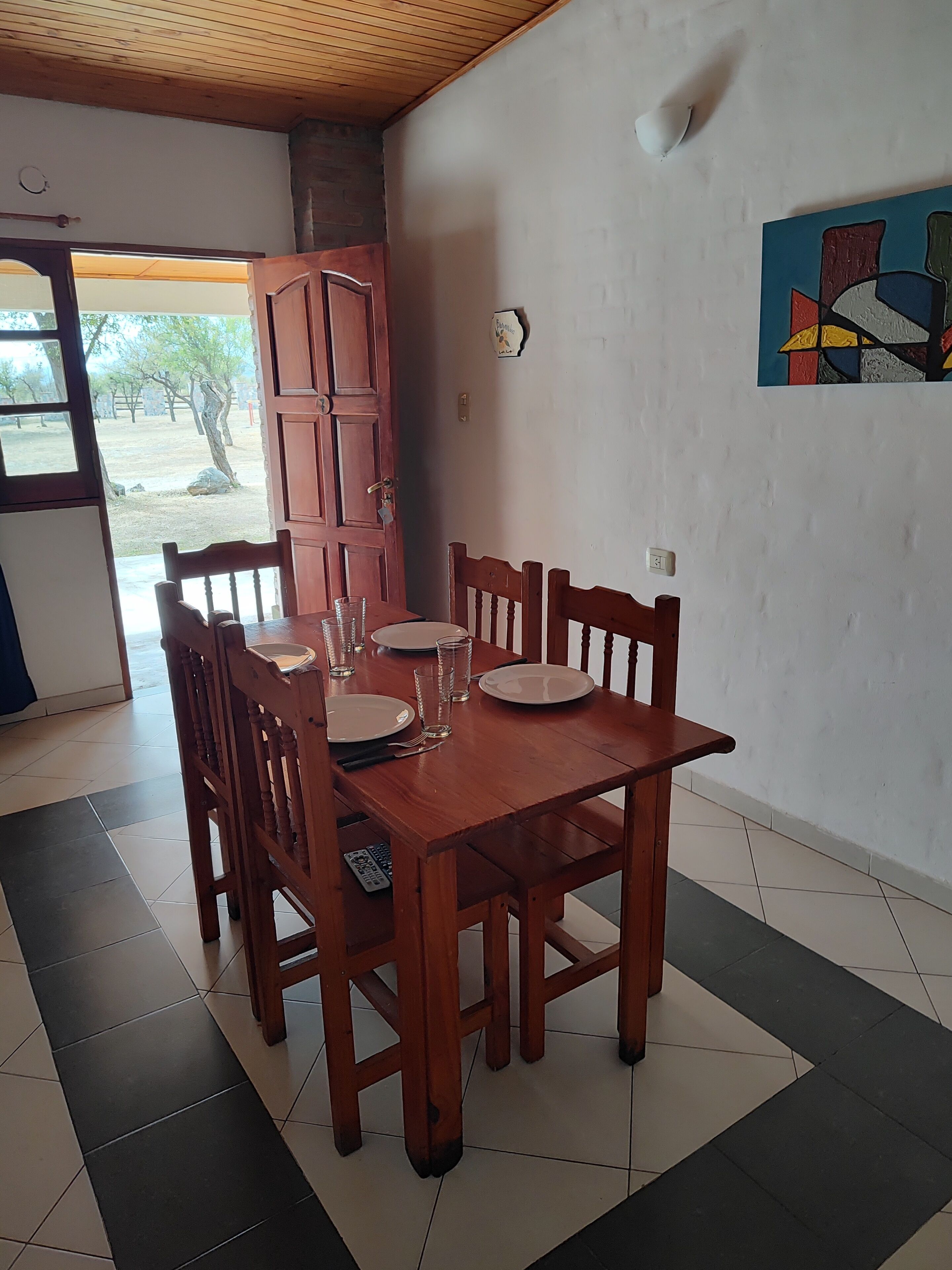 Un comedor con una mesa de madera dispuesta para cuatro personas, rodeada de sillas. Una puerta da al exterior y hay un cuadro en la pared.