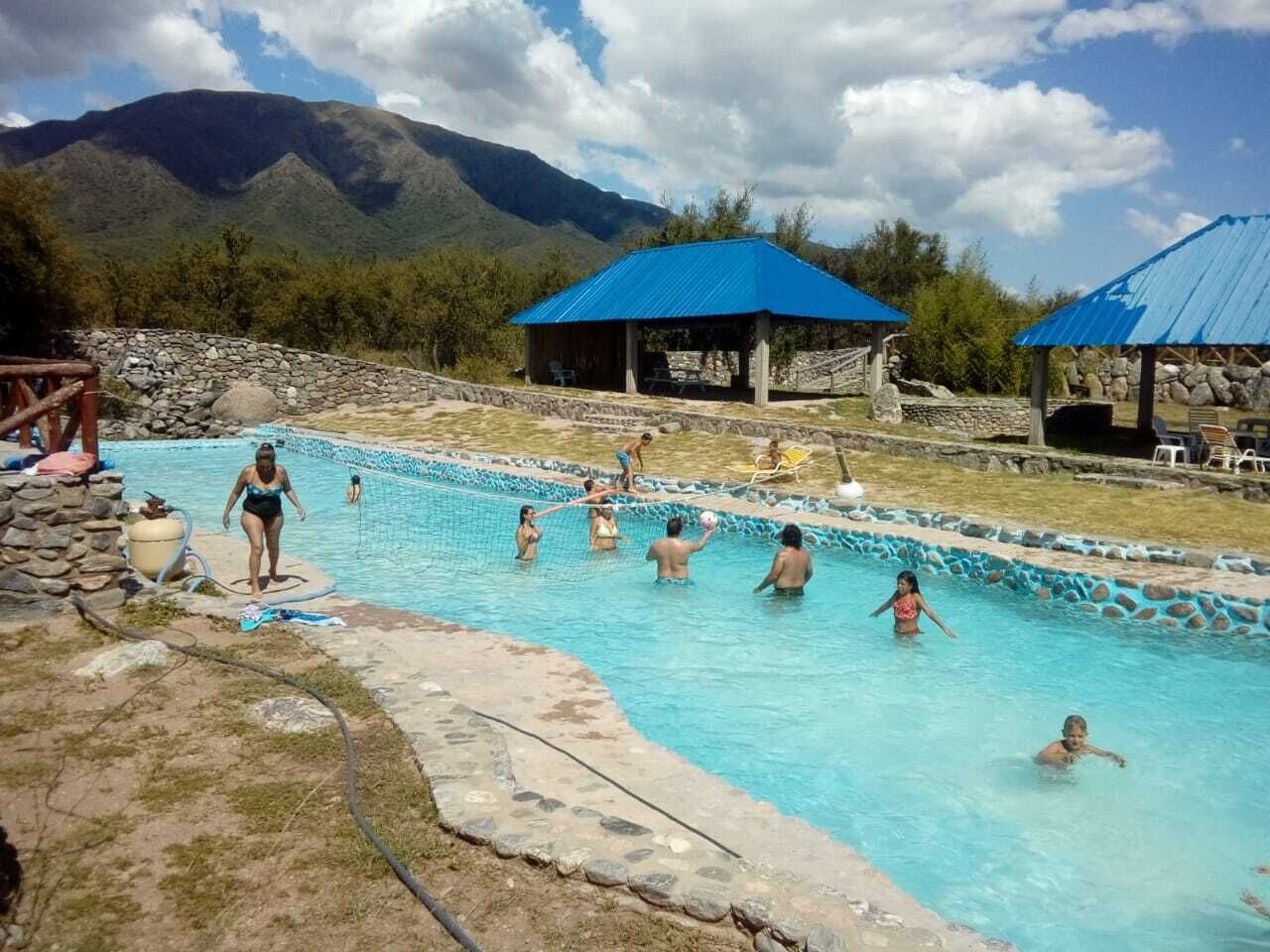 Una piscina con gente jugando, rodeada de muros de piedra y un paisaje montañoso.