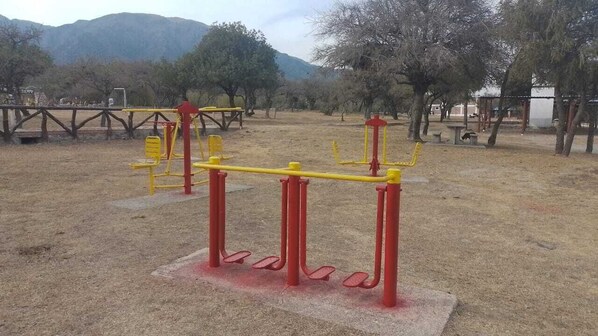 An outdoor exercise area with equipment, including a seesaw and a balance beam, surrounded by trees and a mountain in the background. - COMPLEJOLAGUNAAZUL (Los Molles)