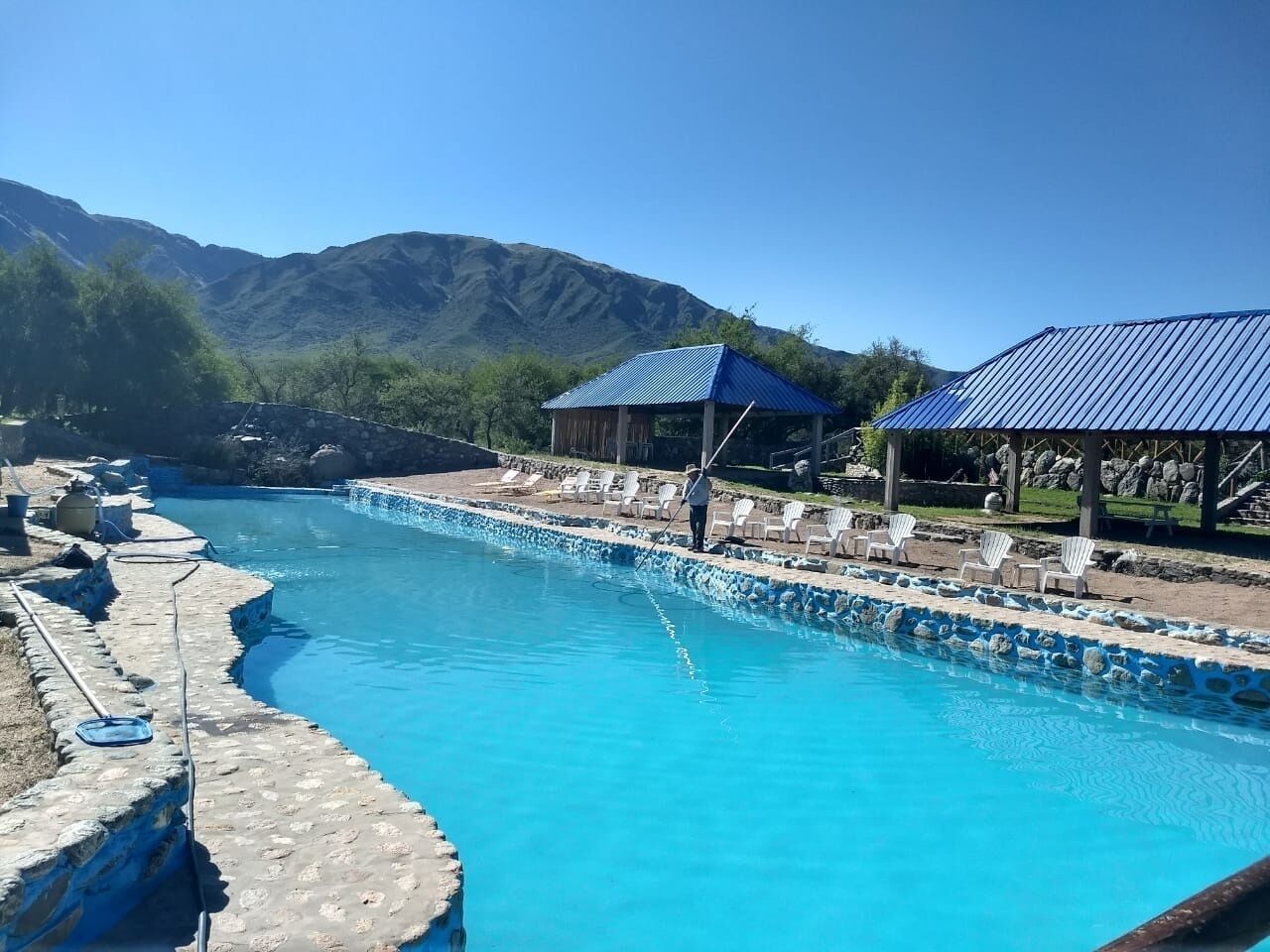 A swimming pool with a stone edge, surrounded by lounge chairs and a covered area with a blue roof.