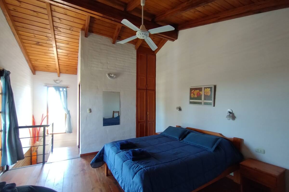 Un dormitorio con techo de madera, una cama con ropa de cama azul, una mesita de noche pequeña, un espejo fijado a la pared y un ventilador de techo.