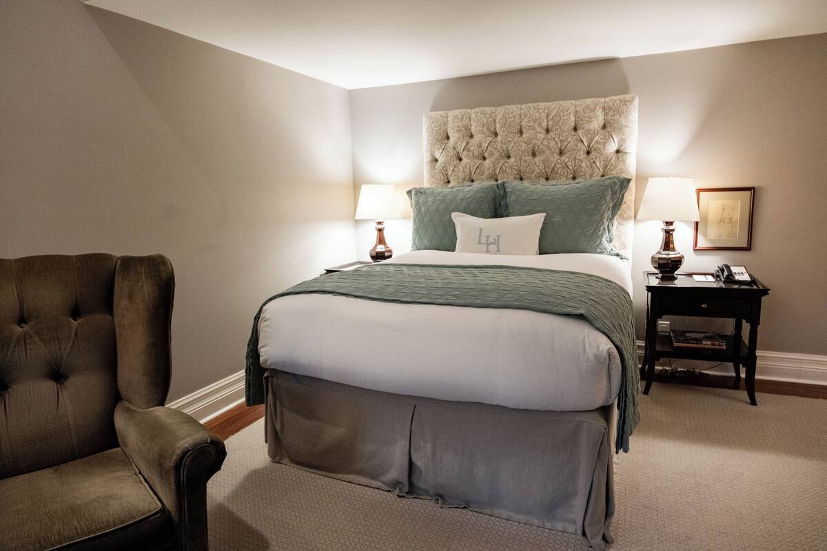 A bedroom with a tufted headboard, a large bed with green bedding, a brown armchair, and a bedside table with a lamp and a framed picture.