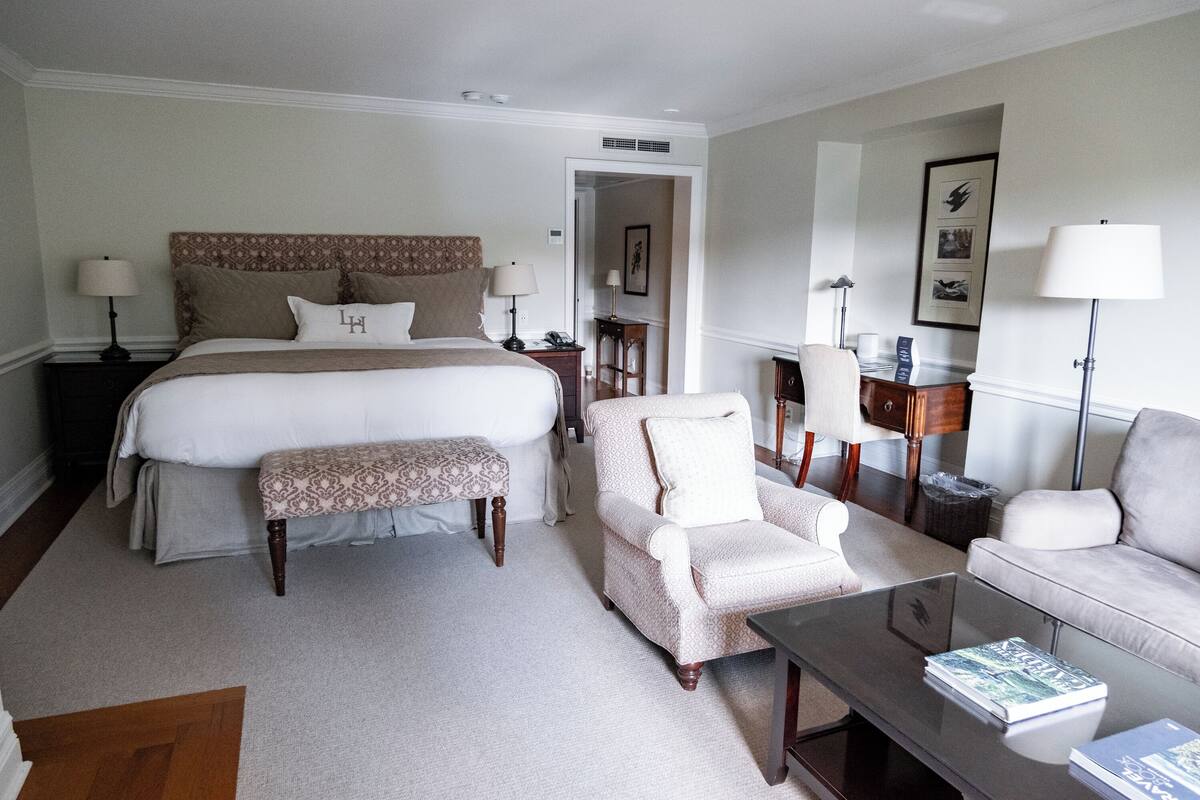 A hotel room with a large bed, a sitting area with a chair and a sofa, a small table, and a bedside table with a lamp.