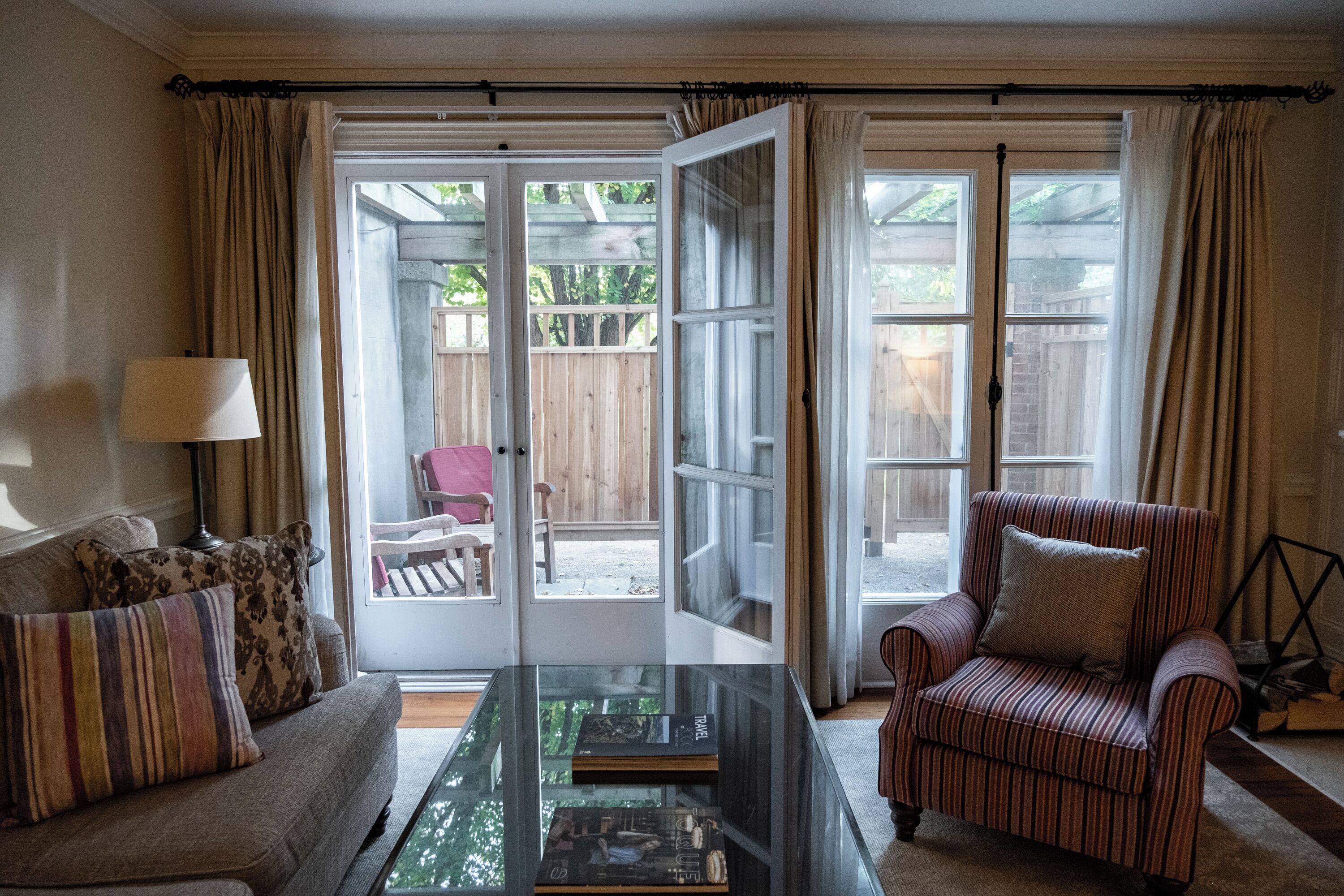 A living room with a sofa, armchair, and coffee table. There is a glass door leading to an outdoor area with a wooden deck and a pink chair.