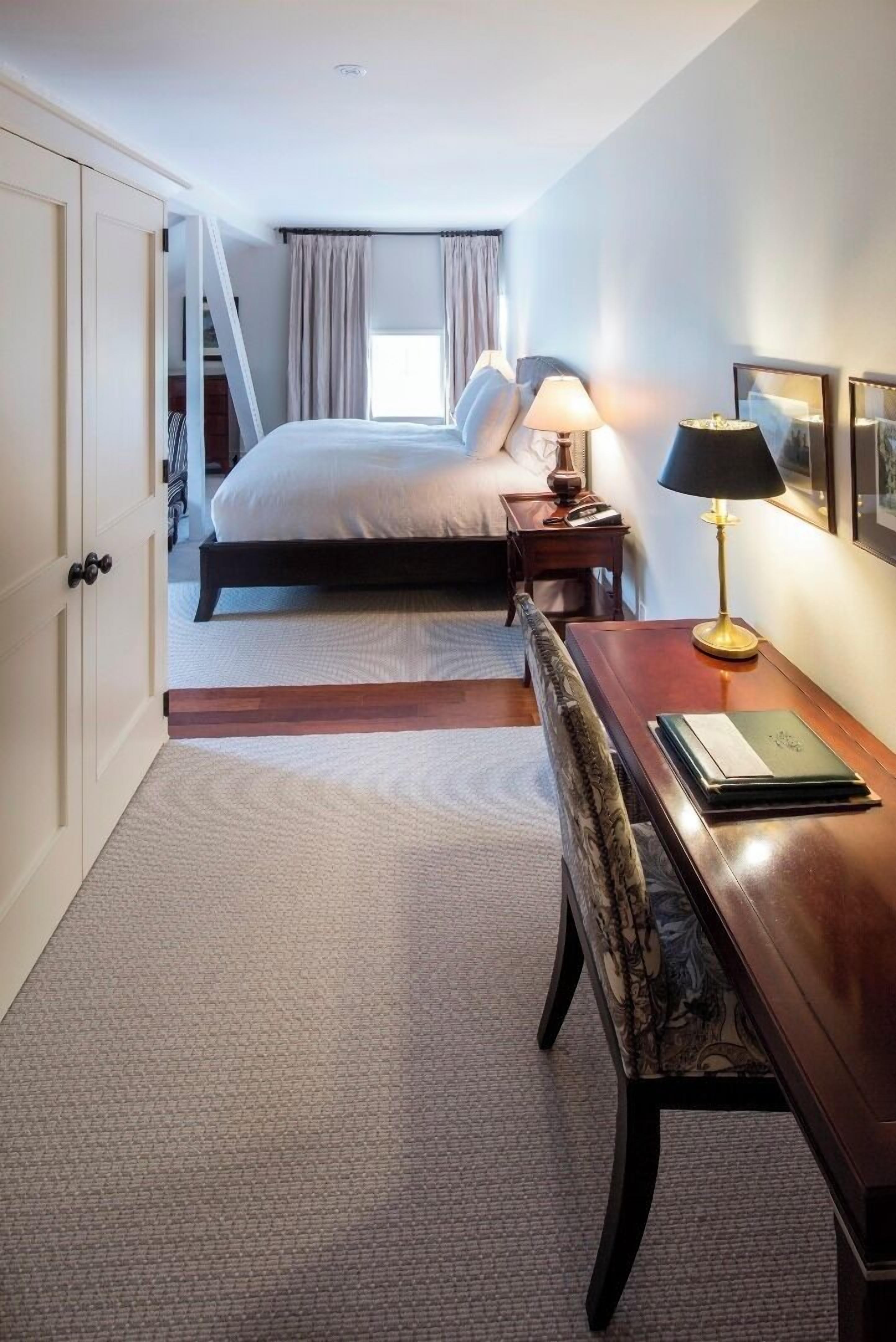 A hotel room with a bed, bedside table, lamp, chair, and a desk with a book.