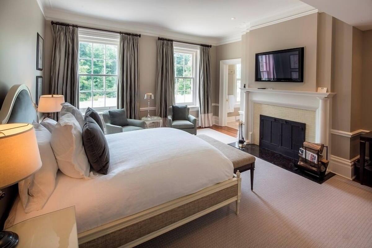 A spacious bedroom with a large bed, a television mounted above a fireplace, and two windows with curtains.