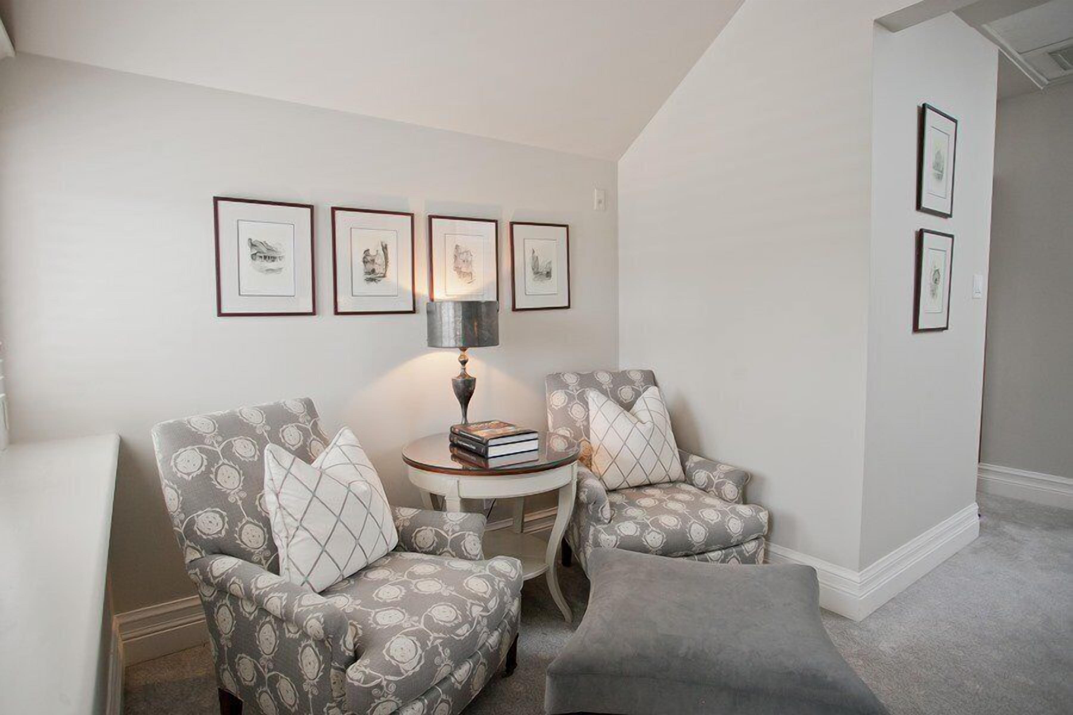 A cozy living room with two patterned armchairs, a small round table with books, and framed artwork on the wall.