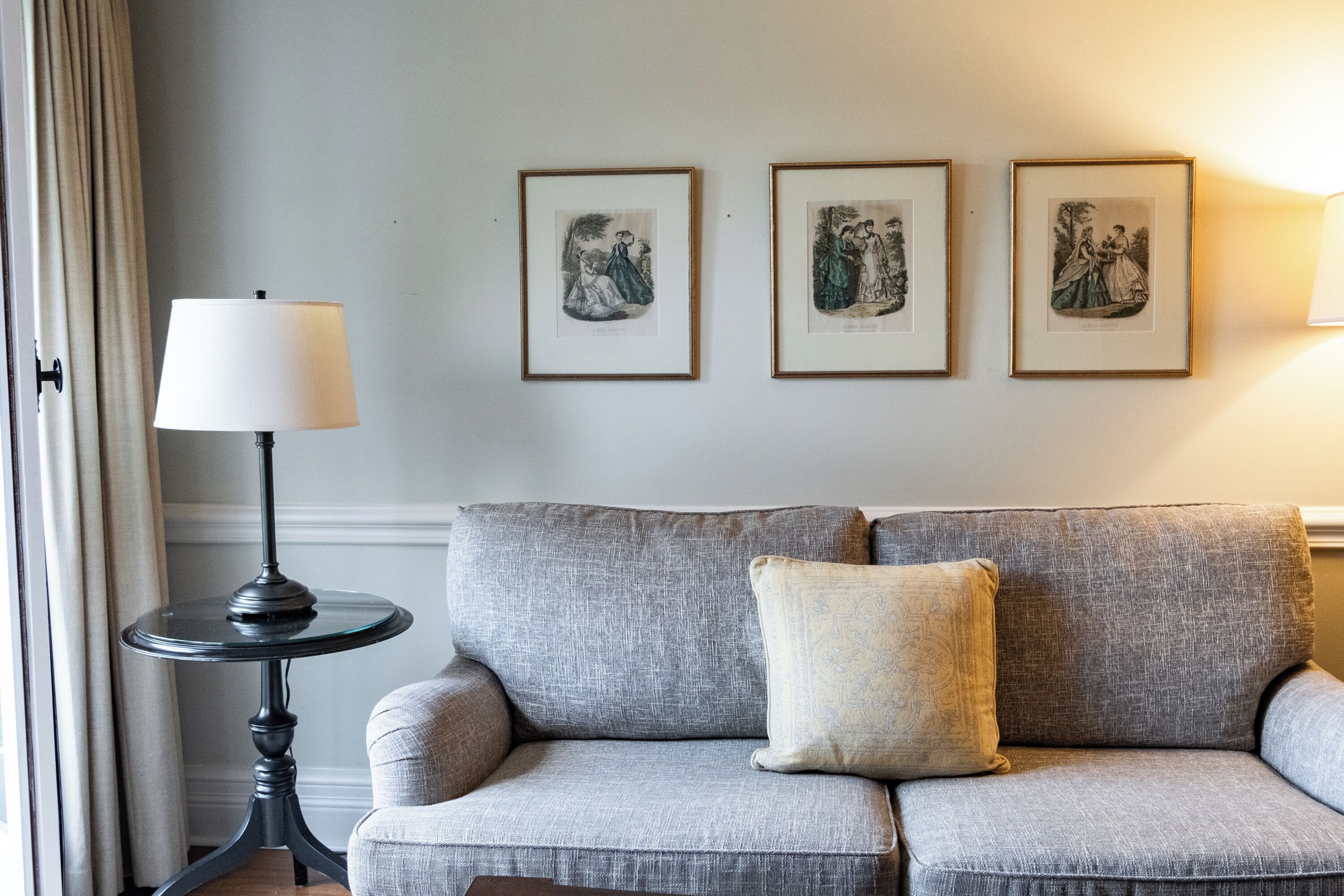 A grey sofa with two cushions, a small round table with a lamp, and three framed pictures on the wall.