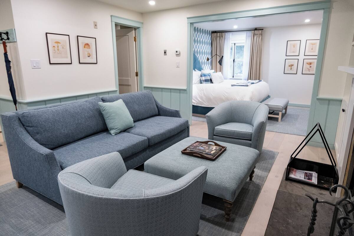A modern hotel room with a grey sofa, a matching armchair, a small ottoman, and a bed with a blue and white patterned headboard.