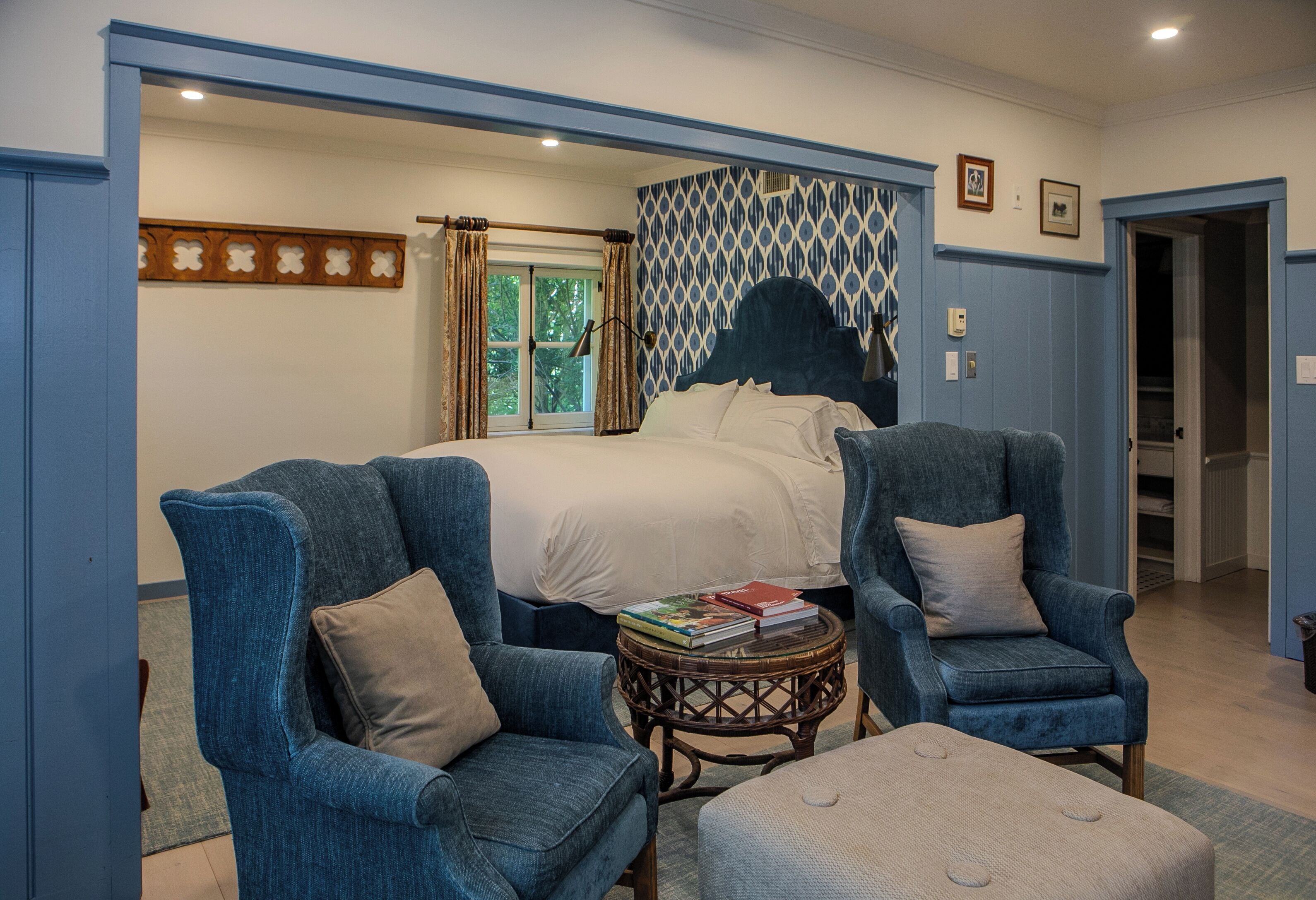 Un dormitorio con una cama, dos sillones, una mesita redonda con libros y un ventanal con cortinas.