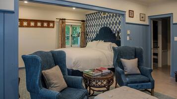 Un dormitorio con una cama, dos sillones, una mesita redonda con libros y un ventanal con cortinas.