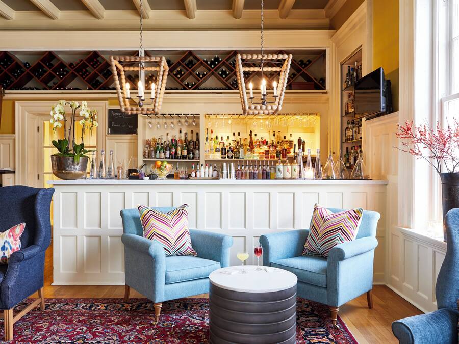 Een bar met twee blauwe fauteuils, een ronde tafel met een glas rode wijn, en een wijnrek boven de bar.