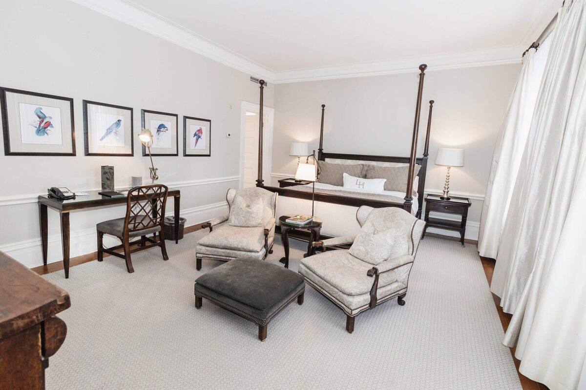 A spacious room with a large bed, two armchairs, a small table, and a desk. There are framed pictures on the wall and a large window with curtains.
