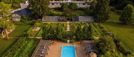 Una piscina grande y rectangular rodeada de sillas de descanso y sombrillas, con un jardín cuidado y una casa grande al fondo.