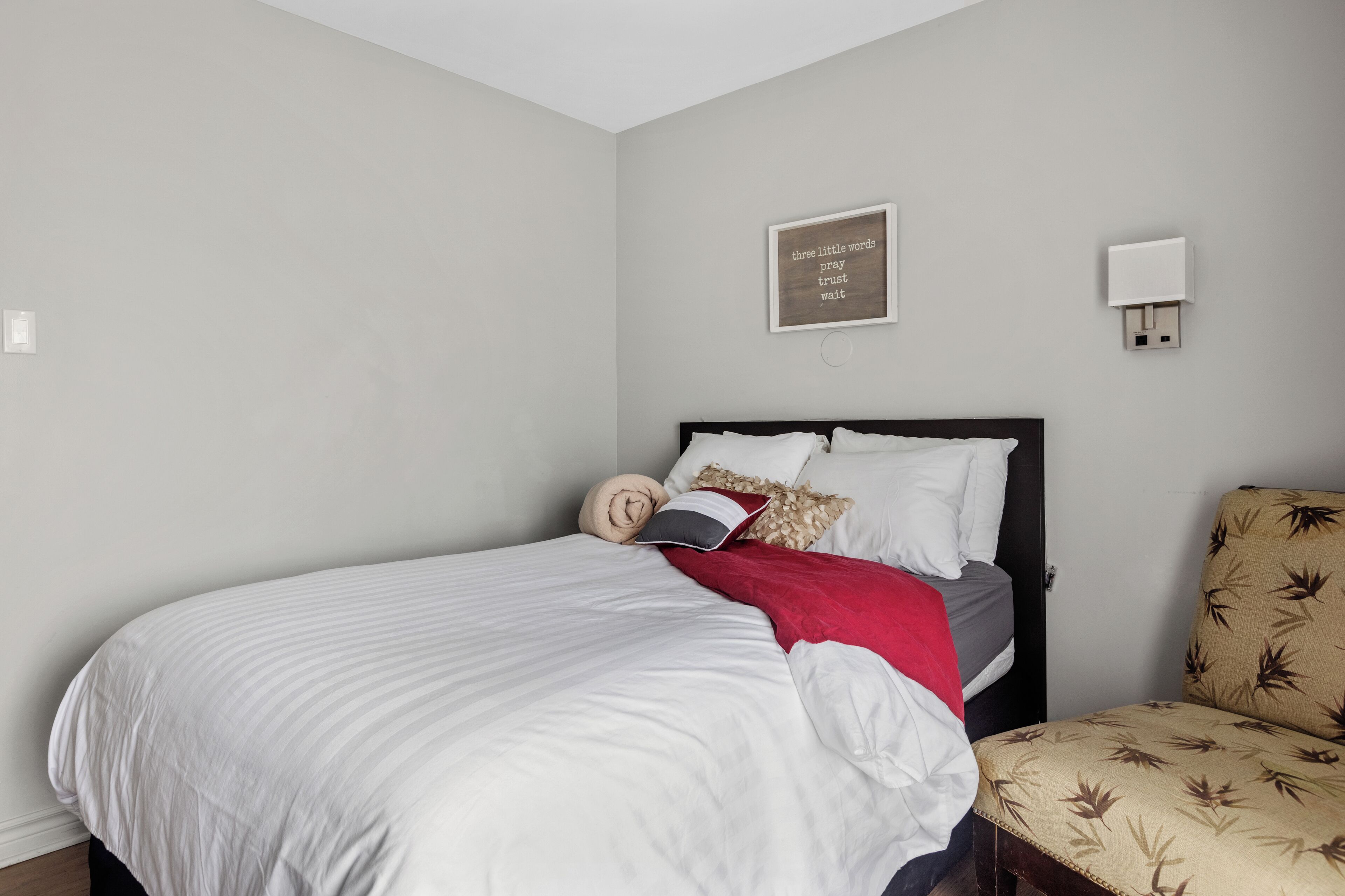 A neatly made bed with white linens, a red throw blanket, and decorative pillows. A wall-mounted light fixture and a framed inspirational quote are visible.