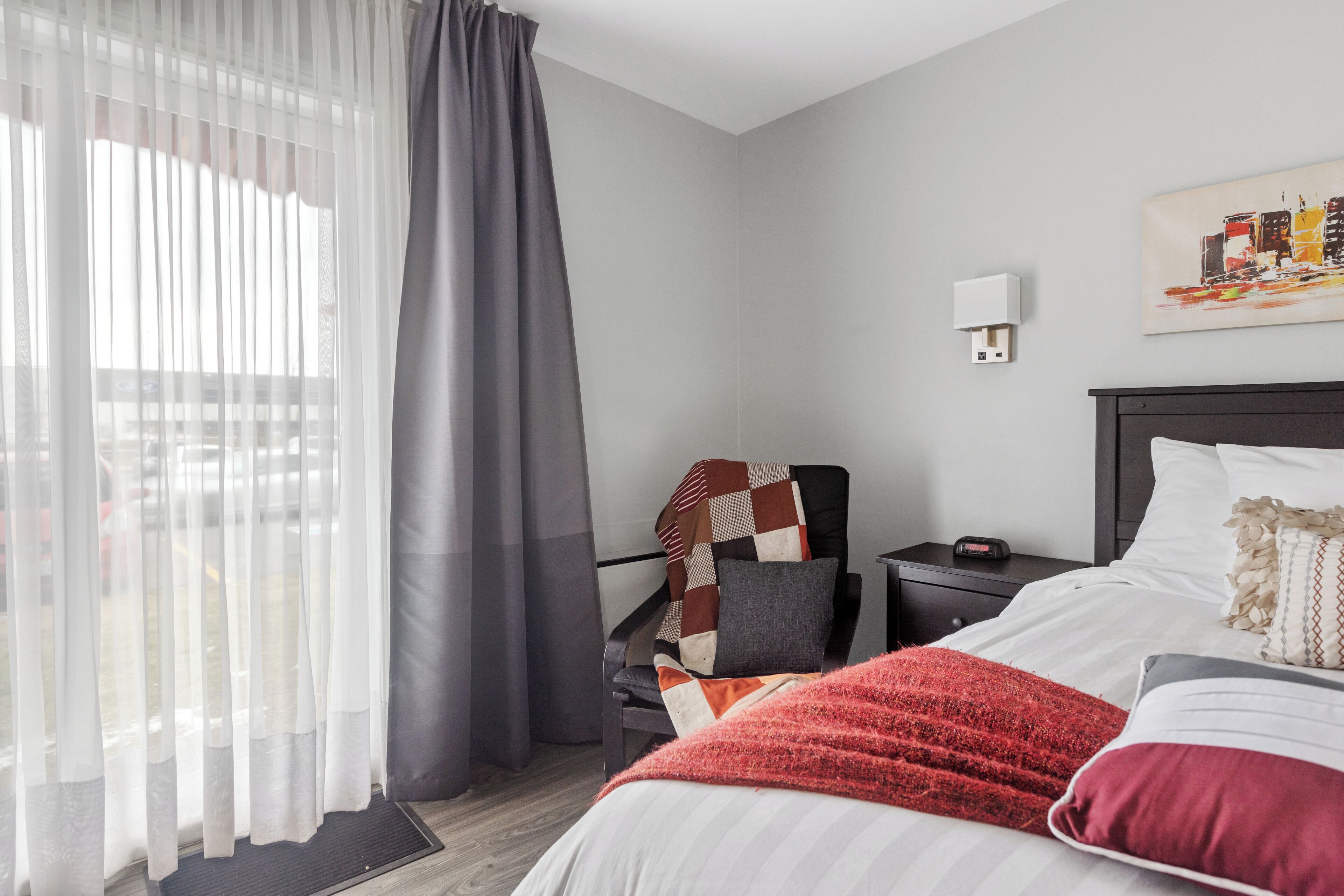 A modern hotel room with a bed, a nightstand, a chair with a quilt, a window with sheer curtains, and a wall-mounted light.