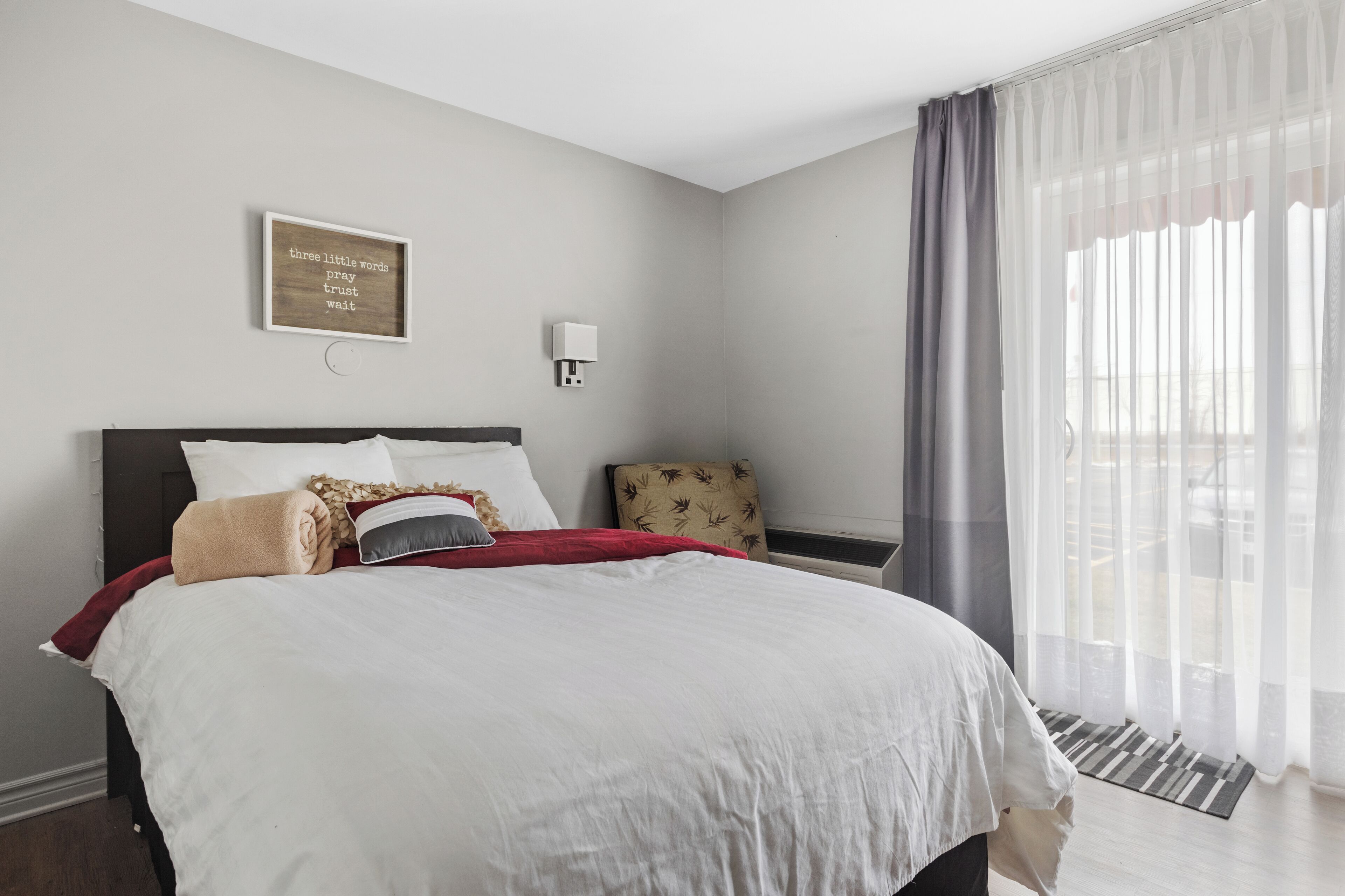 A neatly made bed with a red throw blanket, a wall-mounted light, and a sign with three words.