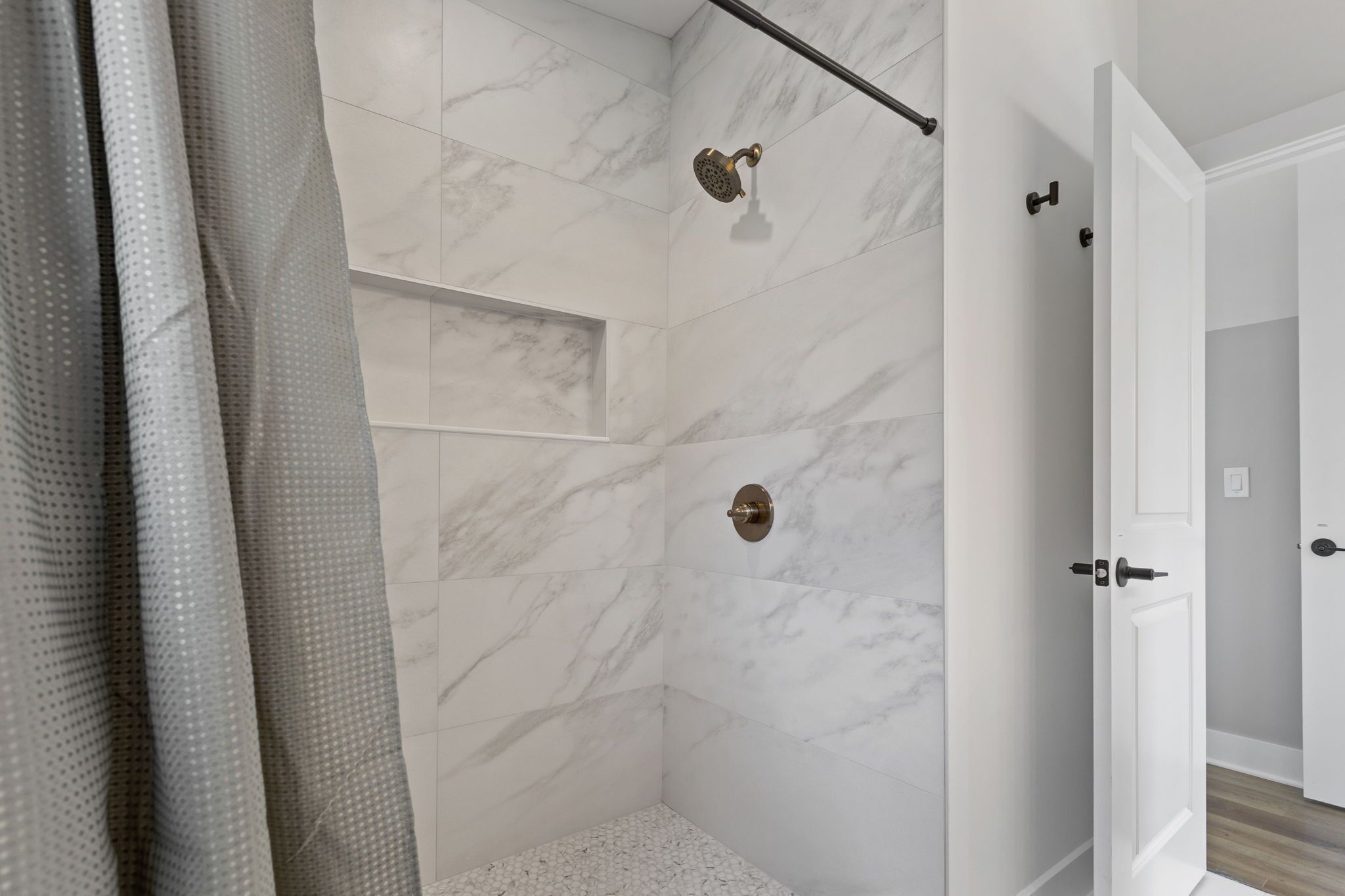 Un bany modern amb parets de marbre blanc, dutxa amb plat de roba i una porta que dona a una altra habitació.