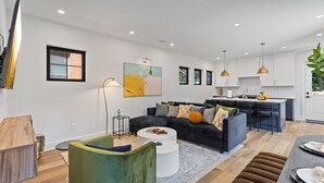 A modern living room with a dark sofa, a green armchair, and a white coffee table. There is a kitchen area with white cabinets and a bar counter. - Rooftop Deck, Hot Tub, Speakeasy, Columbus Luxury (Columbus)
