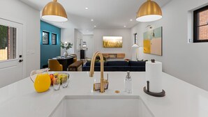 A modern kitchen with a white countertop, gold pendant lights, and a sink. There is a fruit bowl, a pitcher of orange juice, and a bottle of water on the counter. - Rooftop Deck, Hot Tub, Speakeasy, Columbus Luxury (Columbus)