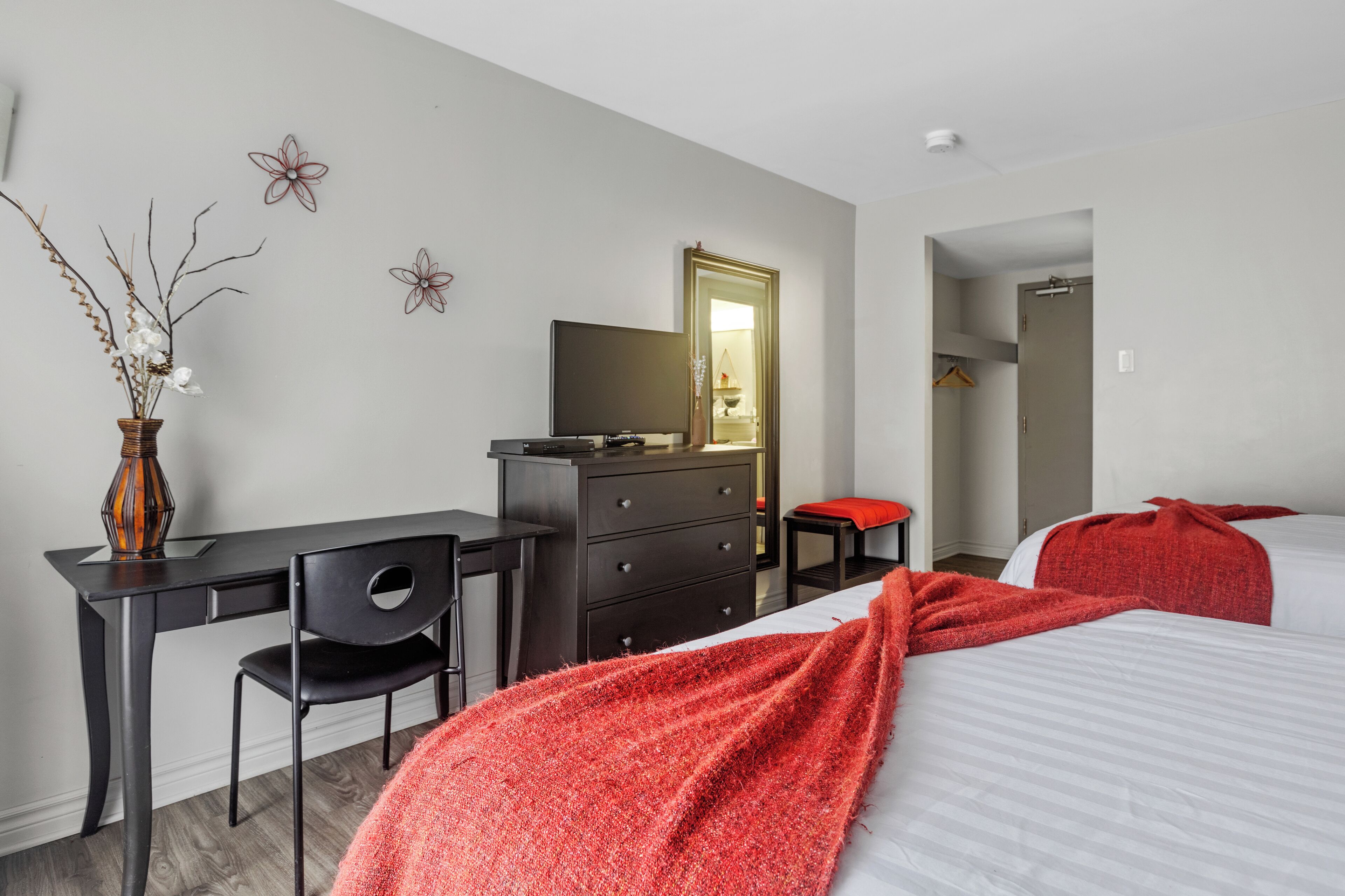 A hotel room with a bed, a desk, a chair, a dresser, a mirror, and a vase with branches.