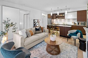 Ein modernes Wohnzimmer mit einer Couch, einem Couchtisch und Sesseln. Es gibt eine Küchenzeile mit Schränken und einer Mikrowelle.