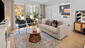 A modern living room with a sofa, coffee table, and chairs. There is a TV, a fireplace, and a large window with a view of greenery. - Luxe King Bed and Private BBQ Terrace (Victoria North)
