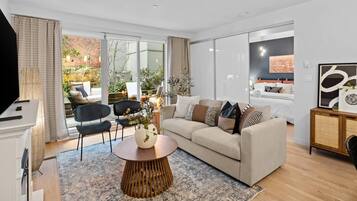 A modern living room with a sofa, coffee table, and chairs. There is a TV, a fireplace, and a large window with a view of greenery.