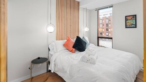 A modern hotel room with a large bed, a bedside table, a round floor lamp, and a view of the city through a large window. - Waterfront Stay with Balcony and BBQ (Victoria Central British Columbia Provincial Government)