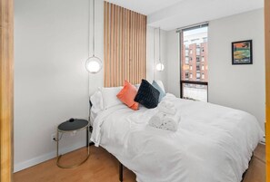 A modern hotel room with a large bed, a bedside table, a round floor lamp, and a view of the city through a large window. - Waterfront Stay with Balcony and BBQ (Victoria Central British Columbia Provincial Government)