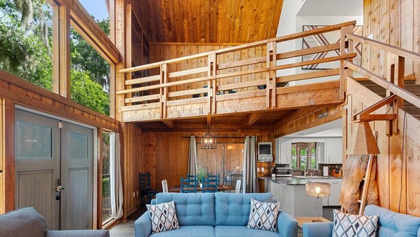 Amplia sala de estar con un juego de sofá azul, una escalera de madera y una cocina al fondo.