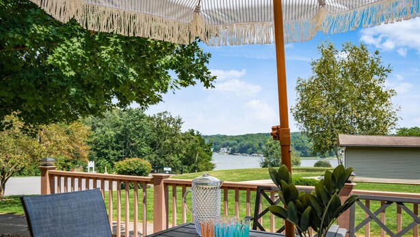 A wooden deck with a table and chairs, overlooking a lake with trees and a building in the background.